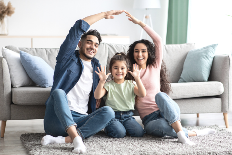 A smiling family of three sits on a rug in front of a gray sofa. The adults make a roof shape with their arms over the child, who holds up both hands and smiles at the camera.